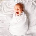 Słodki noworodek ziewający owinięty w przytulny biały otulacz muślinowy, leżący na białej pościeli